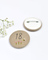 18th Birthday badge. Round badge covered in beige natural linen fabric with the number 18 sewn in black thread using freehand machine embroidery. Little flowers and a tiny bumble bee are delicately hand sewn under the embroidered number. Size 45mm. Handmade by Stitch Galore