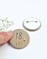 18th Birthday badge. Round badge covered in beige natural linen fabric with the number 18 sewn in black thread using freehand machine embroidery. Little flowers and a tiny bumble bee are delicately hand sewn under the embroidered number. Size 45mm. Handmade by Stitch Galore