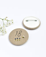 18th Birthday badge. Round badge covered in beige natural linen fabric with the number 18 sewn in black thread using freehand machine embroidery. Little flowers and a tiny bumble bee are delicately hand sewn under the embroidered number. Size 45mm. Handmade by Stitch Galore