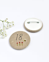 18th Birthday badge. Round badge covered in beige natural linen fabric with the number 18 sewn in black thread using freehand machine embroidery. Little flowers and a tiny bumble bee are delicately hand sewn under the embroidered number. Size 45mm. Handmade by Stitch Galore