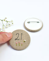 21st Birthday badge. Round badge covered in beige natural linen fabric with the number 21 sewn in black thread using freehand machine embroidery. Little flowers and a tiny bumble bee are delicately hand sewn under the embroidered number. Size 45mm. Handmade by Stitch Galore