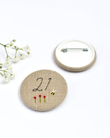 21st Birthday badge. Round badge covered in beige natural linen fabric with the number 21 sewn in black thread using freehand machine embroidery. Little flowers and a tiny bumble bee are delicately hand sewn under the embroidered number. Size 45mm. Handmade by Stitch Galore