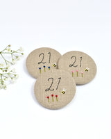 21st Birthday badge. Round badge covered in beige natural linen fabric with the number 21 sewn in black thread using freehand machine embroidery. Little flowers and a tiny bumble bee are delicately hand sewn under the embroidered number. Size 45mm. Handmade by Stitch Galore