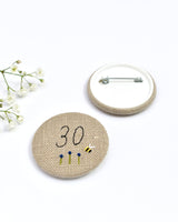 30th Birthday badge. Round badge covered in beige natural linen fabric with the number 30 sewn in black thread using freehand machine embroidery. Little flowers and a tiny bumble bee are delicately hand sewn under the embroidered number. Size 45mm. Handmade by Stitch Galore