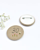 30th Birthday badge. Round badge covered in beige natural linen fabric with the number 30 sewn in black thread using freehand machine embroidery. Little flowers and a tiny bumble bee are delicately hand sewn under the embroidered number. Size 45mm. Handmade by Stitch Galore