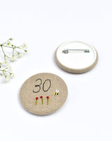 30th Birthday badge. Round badge covered in beige natural linen fabric with the number 30 sewn in black thread using freehand machine embroidery. Little flowers and a tiny bumble bee are delicately hand sewn under the embroidered number. Size 45mm. Handmade by Stitch Galore