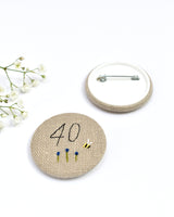 40th Birthday badge. Round badge covered in beige natural linen fabric with the number 40 sewn in black thread using freehand machine embroidery. Little flowers and a tiny bumble bee are delicately hand sewn under the embroidered number. Size 45mm. Handmade by Stitch Galore
