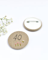 40th Birthday badge. Round badge covered in beige natural linen fabric with the number 40 sewn in black thread using freehand machine embroidery. Little flowers and a tiny bumble bee are delicately hand sewn under the embroidered number. Size 45mm. Handmade by Stitch Galore