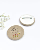 40th Birthday badge. Round badge covered in beige natural linen fabric with the number 40 sewn in black thread using freehand machine embroidery. Little flowers and a tiny bumble bee are delicately hand sewn under the embroidered number. Size 45mm. Handmade by Stitch Galore