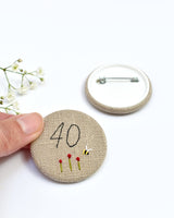 40th Birthday badge. Round badge covered in beige natural linen fabric with the number 40 sewn in black thread using freehand machine embroidery. Little flowers and a tiny bumble bee are delicately hand sewn under the embroidered number. Size 45mm. Handmade by Stitch Galore