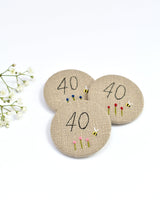 40th Birthday badge. Round badge covered in beige natural linen fabric with the number 40 sewn in black thread using freehand machine embroidery. Little flowers and a tiny bumble bee are delicately hand sewn under the embroidered number. Size 45mm. Handmade by Stitch Galore