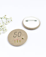 50th Birthday badge. Round badge covered in beige natural linen fabric with the number 50 sewn in black thread using freehand machine embroidery. Little flowers and a tiny bumble bee are delicately hand sewn under the embroidered number. Size 45mm. Handmade by Stitch Galore