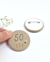 50th Birthday badge. Round badge covered in beige natural linen fabric with the number 50 sewn in black thread using freehand machine embroidery. Little flowers and a tiny bumble bee are delicately hand sewn under the embroidered number. Size 45mm. Handmade by Stitch Galore