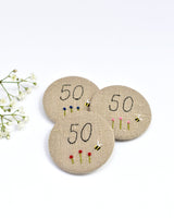 50th Birthday badge. Round badge covered in beige natural linen fabric with the number 50 sewn in black thread using freehand machine embroidery. Little flowers and a tiny bumble bee are delicately hand sewn under the embroidered number. Size 45mm. Handmade by Stitch Galore