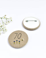 70th Birthday badge. Round badge covered in beige natural linen fabric with the number 70 sewn in black thread using freehand machine embroidery. Little flowers and a tiny bumble bee are delicately hand sewn under the embroidered number. Size 45mm. Handmade by Stitch Galore