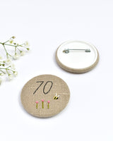 70th Birthday badge. Round badge covered in beige natural linen fabric with the number 70 sewn in black thread using freehand machine embroidery. Little flowers and a tiny bumble bee are delicately hand sewn under the embroidered number. Size 45mm. Handmade by Stitch Galore