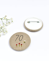 70th Birthday badge. Round badge covered in beige natural linen fabric with the number 70 sewn in black thread using freehand machine embroidery. Little flowers and a tiny bumble bee are delicately hand sewn under the embroidered number. Size 45mm. Handmade by Stitch Galore