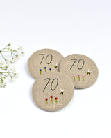 70th Birthday badge. Round badge covered in beige natural linen fabric with the number 70 sewn in black thread using freehand machine embroidery. Little flowers and a tiny bumble bee are delicately hand sewn under the embroidered number. Size 45mm. Handmade by Stitch Galore