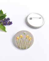 Embroidered daffodil pin badge. Four little yellow daffodil flowers and a tiny bumble bee are delicately hand sewn onto beige natural linen fabric, covering the front of the small round badge.
The round badge measures 45mm in diameter and comes attached to a branded white card.
Handmade by Stitch Galore