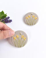 mbroidered daffodil pin badge. Four little yellow daffodil flowers and a tiny bumble bee are delicately hand sewn onto beige natural linen fabric, covering the front of the small round badge.
The round badge measures 45mm in diameter and comes attached to a branded white card.
Handmade by Stitch Galore