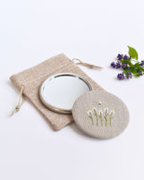Embroidered daisy pocket mirror. Little white daisy flowers and a tiny bumble bee are delicately hand sewn onto natural linen fabric, covering the back of the small round hand mirror.
The round pocket mirror measures 77mm in diameter and comes with a linen drawstring pouch.
Image shows the pretty pocket mirror sitting on top of the linen pouch.