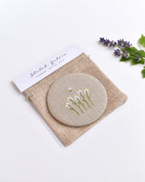 Embroidered daisy pocket mirror. Little white daisy flowers and a tiny bumble bee are delicately hand sewn onto natural linen fabric, covering the back of the small round hand mirror.
The round pocket mirror measures 77mm in diameter and comes with a linen drawstring pouch.
Image shows the pretty pocket mirror sitting on top of the linen pouch.