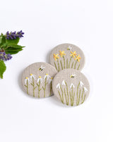 Embroidered floral pin badge. Little flowers and a tiny bumble bee are delicately hand sewn onto beige natural linen fabric, covering the front of the small round badges.
The round badge measures 45mm in diameter and comes attached to a branded white card.
Handmade by Stitch Galore
