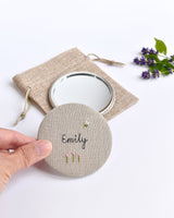 Embroidered personalised pocket mirror. An embroidered name, little flowers and a tiny bumble bee are delicately sewn onto natural linen fabric, covering the back of the small round hand mirror.
The round pocket mirror measures 77mm in diameter and comes with a linen drawstring pouch.
Image shows the pretty pocket mirror sitting on top of the linen pouch.
Handmade by Stitch Galore.