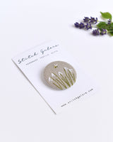 Embroidered snowdrop pin badge. Five little white snowdrop flowers and a tiny bumble bee are delicately hand sewn onto beige natural linen fabric, covering the front of the small round badge.
The round badge measures 45mm in diameter and comes attached to a branded white card.
Handmade by Stitch Galore