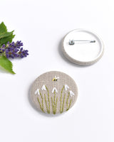 Embroidered snowdrop pin badge. Five little white snowdrop flowers and a tiny bumble bee are delicately hand sewn onto beige natural linen fabric, covering the front of the small round badge.
The round badge measures 45mm in diameter and comes attached to a branded white card.
Handmade by Stitch Galore
