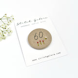 18th Birthday badge. Round badge covered in beige natural linen fabric with the number 18 sewn in black thread using freehand machine embroidery. Little flowers and a tiny bumble bee are delicately hand sewn under the embroidered number. Size 45mm. Handmade by Stitch Galore