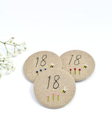 18th Birthday badge. Round badge covered in beige natural linen fabric with the number 18 sewn in black thread using freehand machine embroidery. Little flowers and a tiny bumble bee are delicately hand sewn under the embroidered number. Size 45mm. Handmade by Stitch Galore