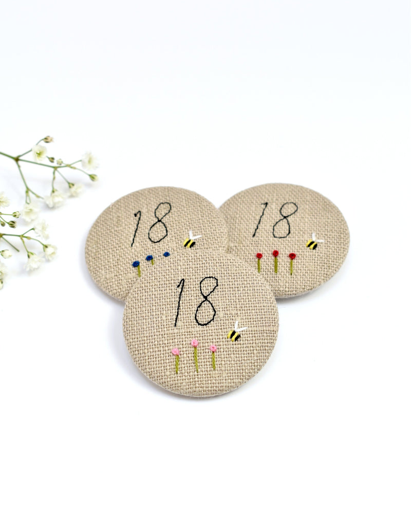 18th Birthday badge. Round badge covered in beige natural linen fabric with the number 18 sewn in black thread using freehand machine embroidery. Little flowers and a tiny bumble bee are delicately hand sewn under the embroidered number. Size 45mm. Handmade by Stitch Galore