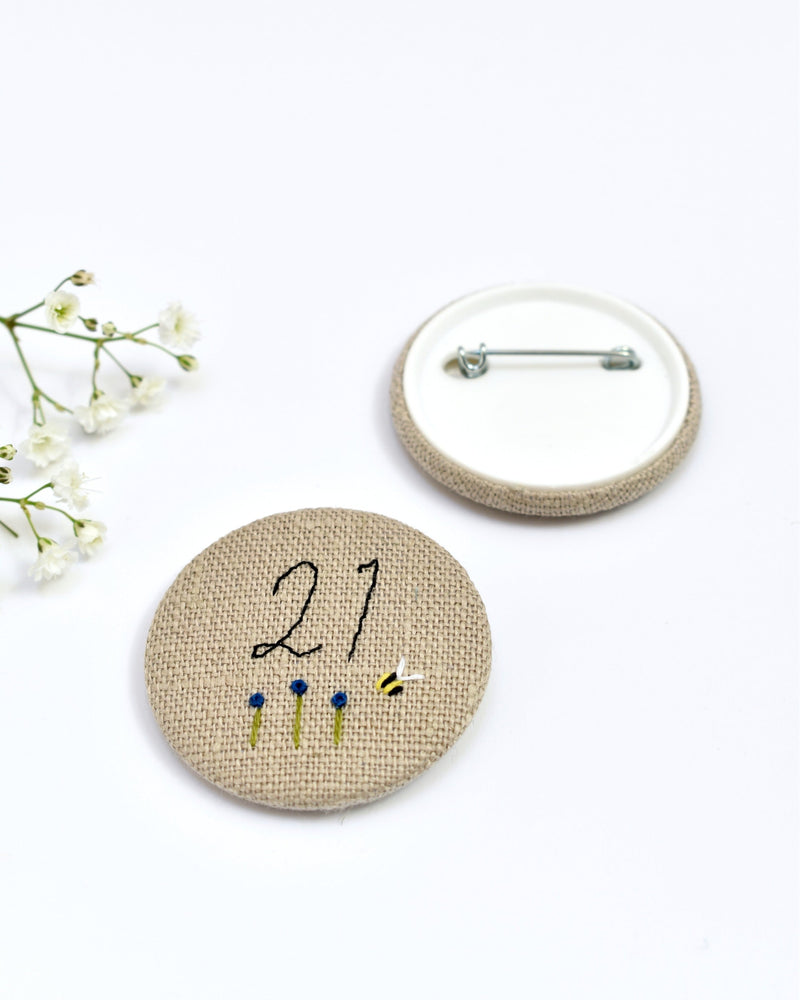 21st Birthday badge. Round badge covered in beige natural linen fabric with the number 21 sewn in black thread using freehand machine embroidery. Little flowers and a tiny bumble bee are delicately hand sewn under the embroidered number. Size 45mm. Handmade by Stitch Galore