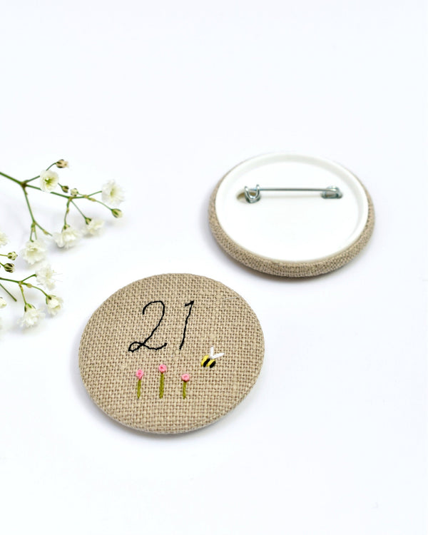 21st Birthday badge. Round badge covered in beige natural linen fabric with the number 21 sewn in black thread using freehand machine embroidery. Little flowers and a tiny bumble bee are delicately hand sewn under the embroidered number. Size 45mm. Handmade by Stitch Galore