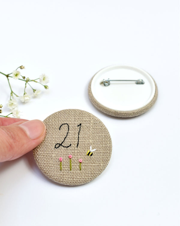 21st Birthday badge. Round badge covered in beige natural linen fabric with the number 21 sewn in black thread using freehand machine embroidery. Little flowers and a tiny bumble bee are delicately hand sewn under the embroidered number. Size 45mm. Handmade by Stitch Galore