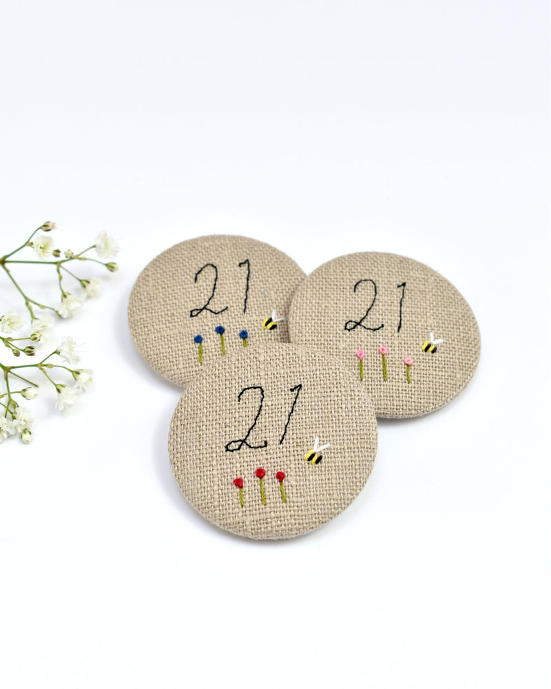 21st Birthday badge. Round badge covered in beige natural linen fabric with the number 21 sewn in black thread using freehand machine embroidery. Little flowers and a tiny bumble bee are delicately hand sewn under the embroidered number. Size 45mm. Handmade by Stitch Galore