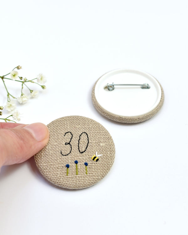 30th Birthday badge. Round badge covered in beige natural linen fabric with the number 30 sewn in black thread using freehand machine embroidery. Little flowers and a tiny bumble bee are delicately hand sewn under the embroidered number. Size 45mm. Handmade by Stitch Galore