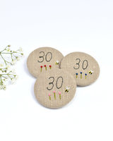 30th Birthday badge. Round badge covered in beige natural linen fabric with the number 30 sewn in black thread using freehand machine embroidery. Little flowers and a tiny bumble bee are delicately hand sewn under the embroidered number. Size 45mm. Handmade by Stitch Galore