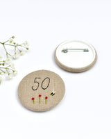 50th Birthday badge. Round badge covered in beige natural linen fabric with the number 50 sewn in black thread using freehand machine embroidery. Little flowers and a tiny bumble bee are delicately hand sewn under the embroidered number. Size 45mm. Handmade by Stitch Galore