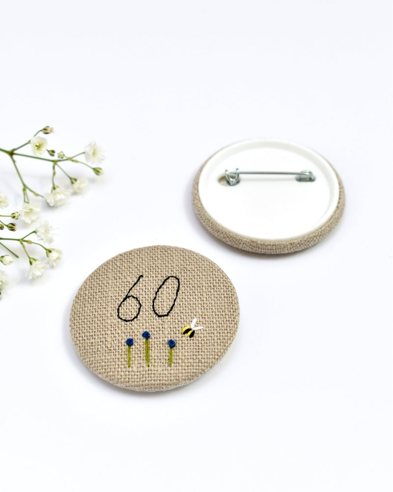 18th Birthday badge. Round badge covered in beige natural linen fabric with the number 18 sewn in black thread using freehand machine embroidery. Little flowers and a tiny bumble bee are delicately hand sewn under the embroidered number. Size 45mm. Handmade by Stitch Galore