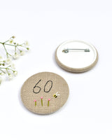 18th Birthday badge. Round badge covered in beige natural linen fabric with the number 18 sewn in black thread using freehand machine embroidery. Little flowers and a tiny bumble bee are delicately hand sewn under the embroidered number. Size 45mm. Handmade by Stitch Galore