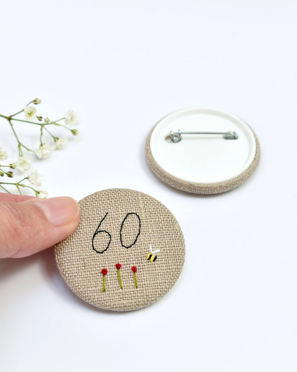 18th Birthday badge. Round badge covered in beige natural linen fabric with the number 18 sewn in black thread using freehand machine embroidery. Little flowers and a tiny bumble bee are delicately hand sewn under the embroidered number. Size 45mm. Handmade by Stitch Galore