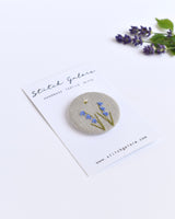 Embroidered bluebell pin badge. |Two little bluebell flowers and a tiny bumble bee are delicately hand sewn onto beige natural linen fabric, covering the front of the small round badge.
The round badge measures 45mm in diameter and comes attached to a branded white card.
Handmade by Stitch Galore