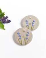 Embroidered bluebell pin badge. |Two little bluebell flowers and a tiny bumble bee are delicately hand sewn onto beige natural linen fabric, covering the front of the small round badge.
The round badge measures 45mm in diameter and comes attached to a branded white card.
Handmade by Stitch Galore
