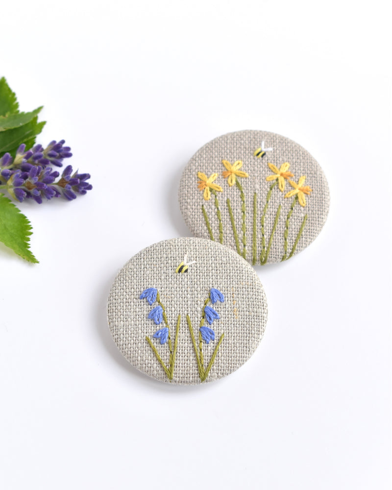 Embroidered bluebell pin badge. |Two little bluebell flowers and a tiny bumble bee are delicately hand sewn onto beige natural linen fabric, covering the front of the small round badge.
The round badge measures 45mm in diameter and comes attached to a branded white card.
Handmade by Stitch Galore