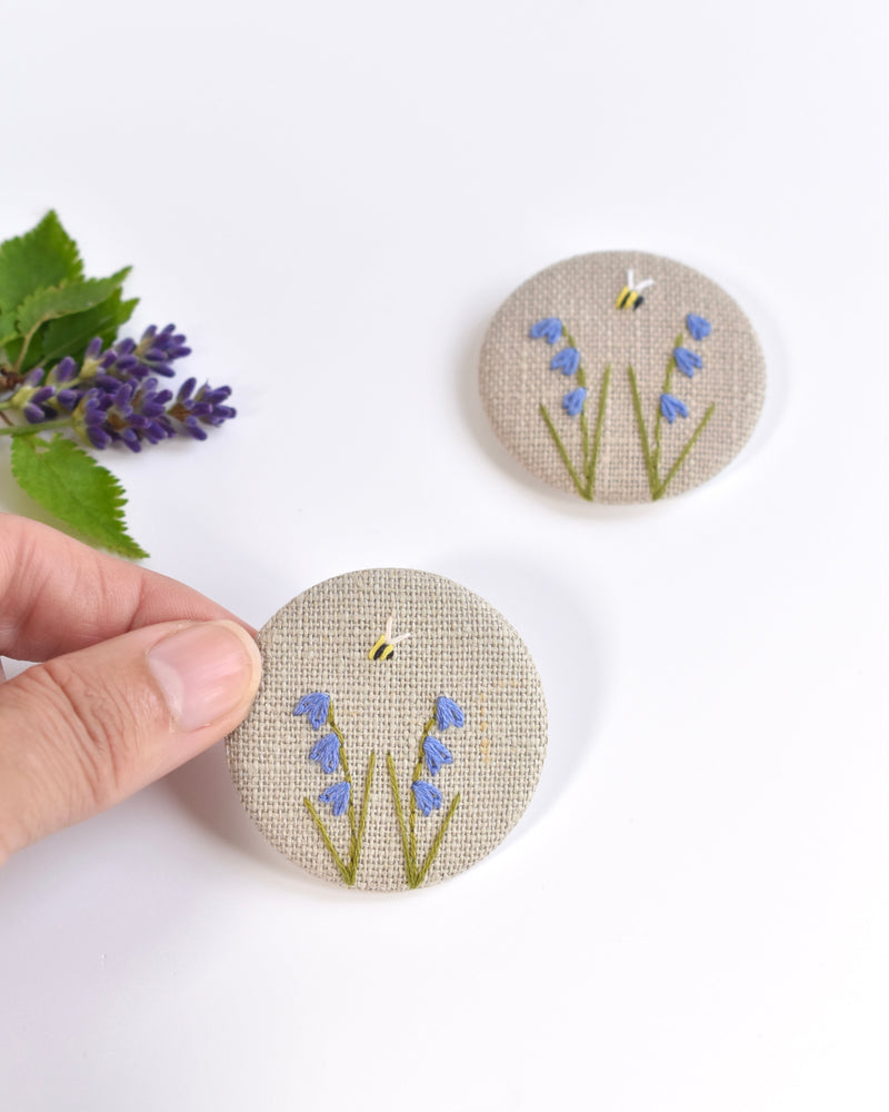 Embroidered bluebell pin badge. |Two little bluebell flowers and a tiny bumble bee are delicately hand sewn onto beige natural linen fabric, covering the front of the small round badge.
The round badge measures 45mm in diameter and comes attached to a branded white card.
Handmade by Stitch Galore