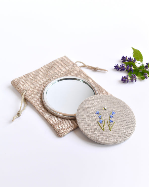 Embroidered bluebell pocket mirror. Little bluebell flowers and a tiny bumble bee are delicately hand sewn onto natural linen fabric, covering the back of the small round hand mirror.
The round pocket mirror measures 77mm in diameter and comes with a linen drawstring pouch.
Image shows the pretty pocket mirror sitting on top of the linen pouch.
