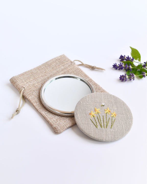 Embroidered daffodil pocket mirror. Little yellow daffodil flowers and a tiny bumble bee are delicately hand sewn onto natural linen fabric, covering the back of the small round hand mirror.
The round pocket mirror measures 77mm in diameter and comes with a linen drawstring pouch.
Image shows the pretty pocket mirror sitting on top of the linen pouch.
