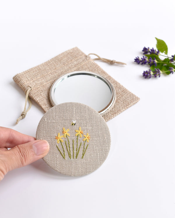Embroidered daffodil pocket mirror. Little yellow daffodil flowers and a tiny bumble bee are delicately hand sewn onto natural linen fabric, covering the back of the small round hand mirror.
The round pocket mirror measures 77mm in diameter and comes with a linen drawstring pouch.
Image shows the pretty pocket mirror being held in a hand.
